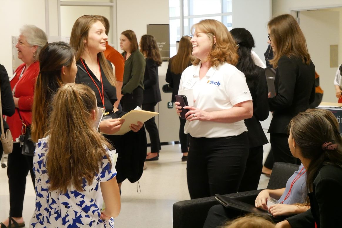 four young women in business dress stand with an older woman in a white company polo, talking