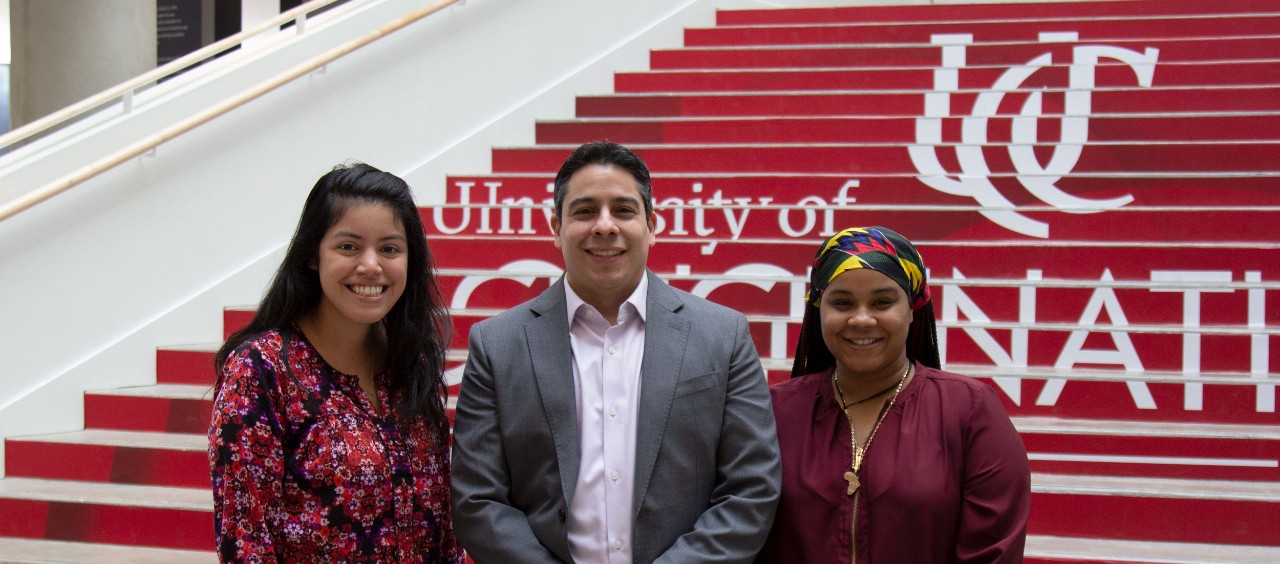 Two women dressed in business casual stand on either side of a man in a suit jacket in front of the staircase at Lindner Hall