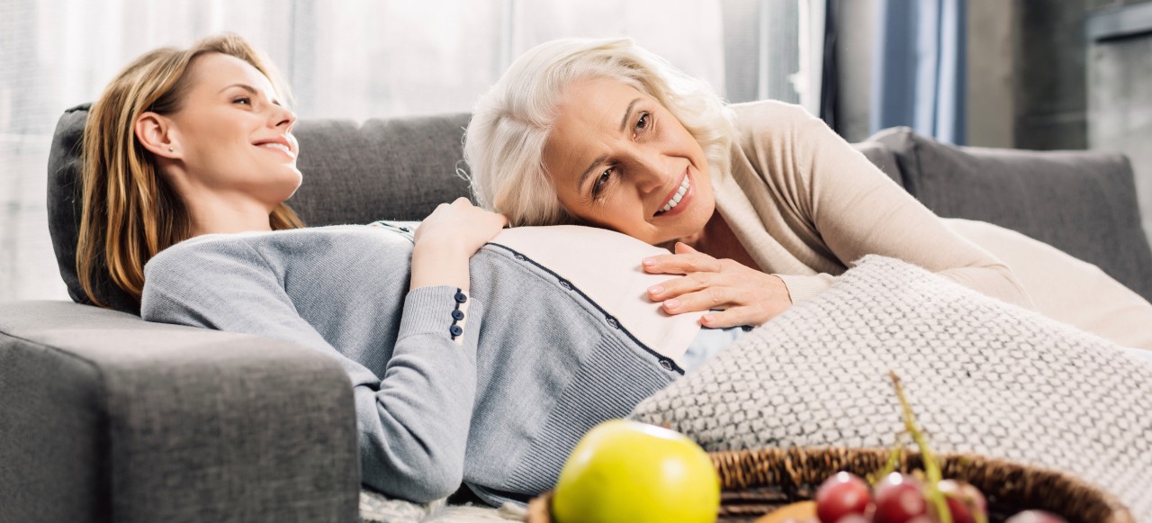 Pregnant woman lies back on couch with her mother leaning on her belly.