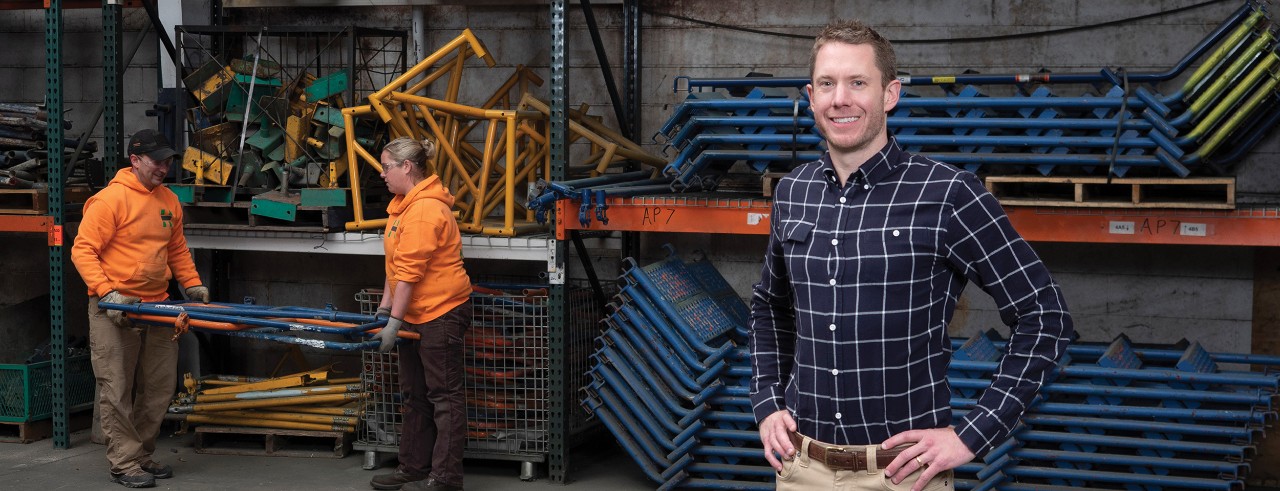 Leadership Development Institute graduate standing in his company's warehouse, with two employees carrying scaffolding in the background.