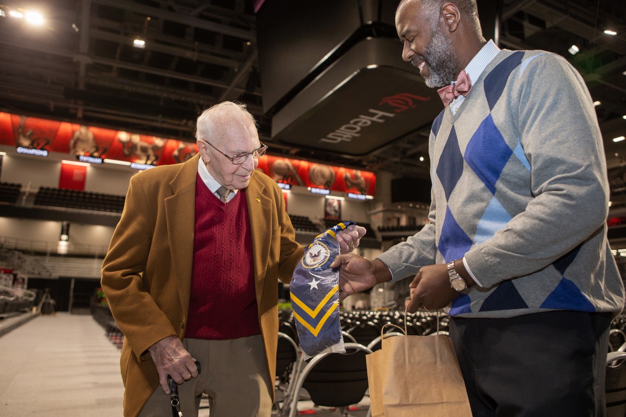Two men standing together, one of whom is handing the other a blue piece of fabric wrapped in plastic. 