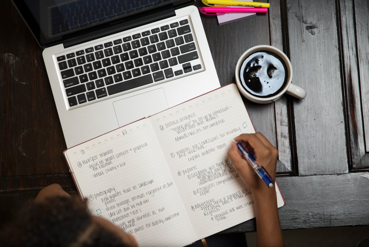A laptop with a note book and someone taking notes