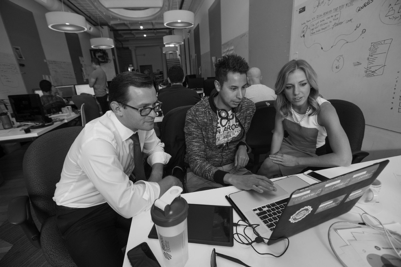 The individuals crowd around a laptop in a bright, innovative-looking office space