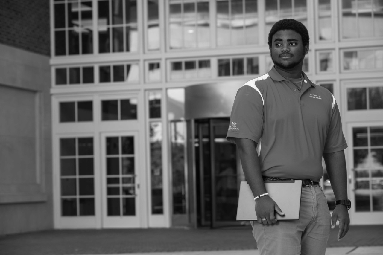 Male student stares past the camera while holding laptop outside campus building
