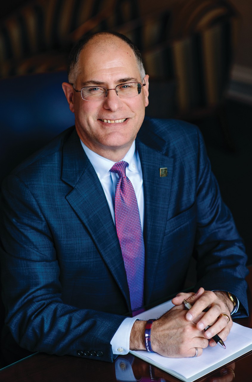 Man in suit sitting at a desk with his hands folded. 