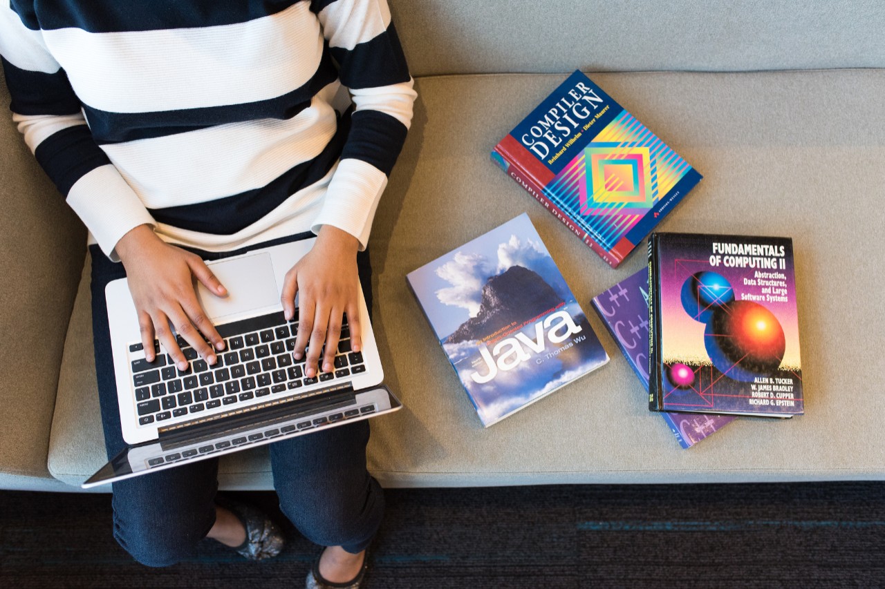 An overhead view of a woman using a laptop computer. Next to her on the couch are a variety of computer programming textbooks.