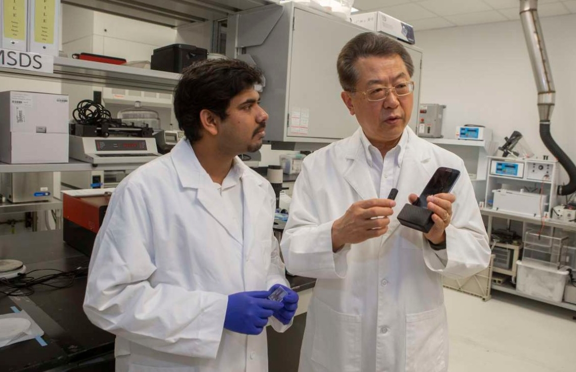 UC professor Chong Ahn and UC student Sthitodhi Ghosh wearing lab coats discuss a smartphone device in an electrical engineering lab.