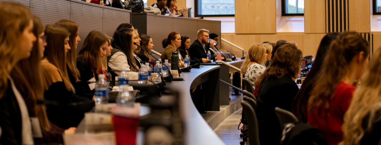 Dozens of young women and young men sit with coffees, waters and notebooks in an auditorium listening and watching what is implied as a speaker at the front of the room