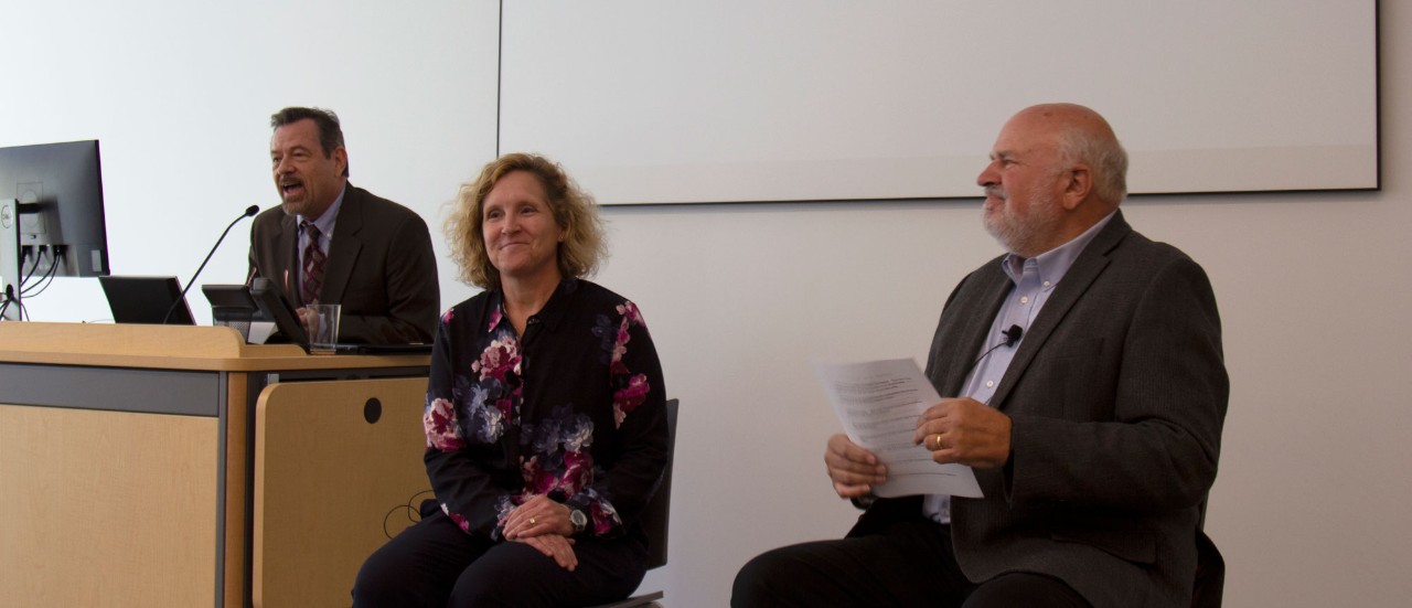 A man in a grey suit in the left in the background stands at a podium and speaks into a microphone, introducing a woman with blonde hair and business professional attire and a man with short hair and a blue shirt and grey jacket in the foreground