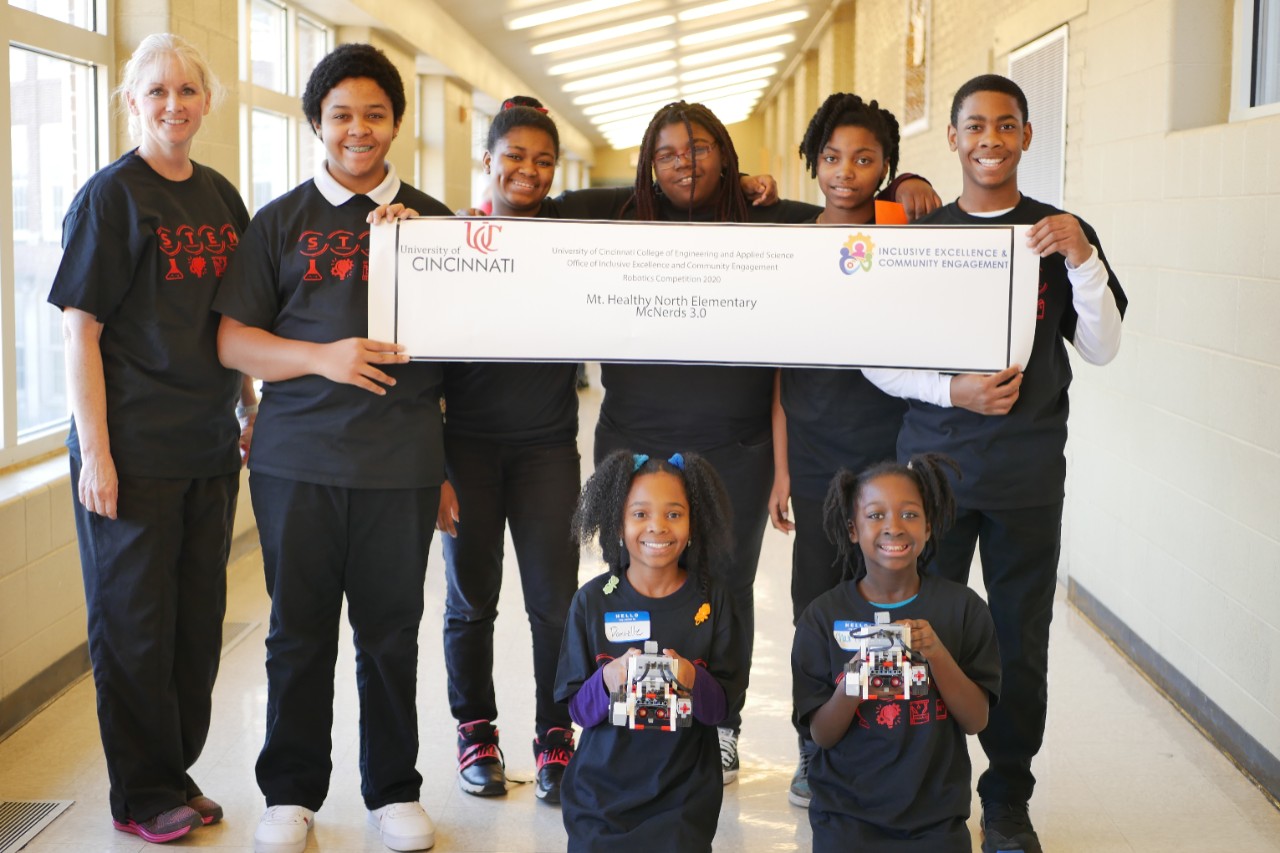 A group of seven elementary aged children and one adult in a bright hallway with a banner that reads their school name, two kneel in front holding Lego robots