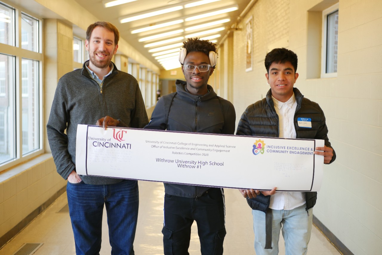 Two high school age boys and one adult male stand with a banner that features their school name