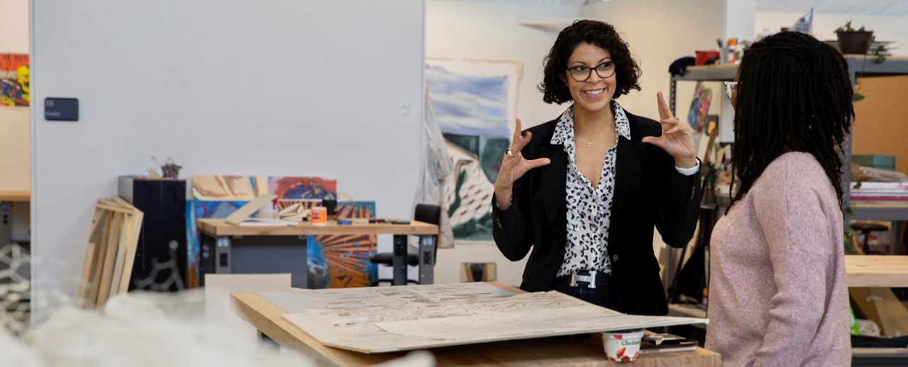 Flavia Bastos stands with her hands in the air teaching a student at a table of art designs 