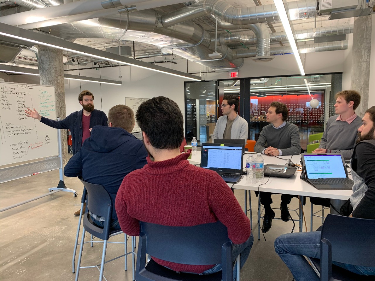 Several students sitting around a table with open laptops watch a presenter speaking and gesturing at a white board