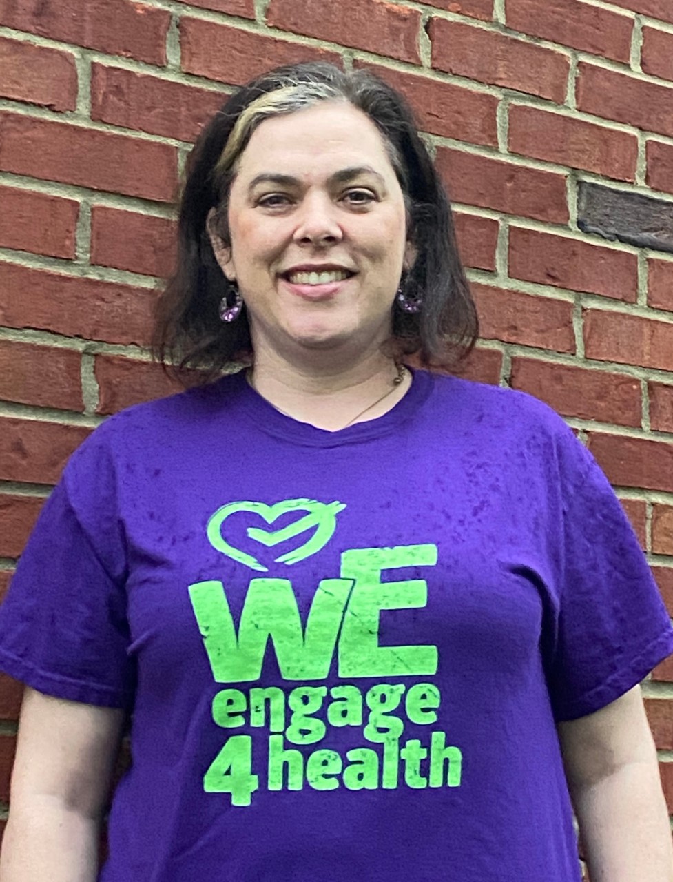 A woman in a We Engage 4 Health t-shirt standing in front of a brick wall