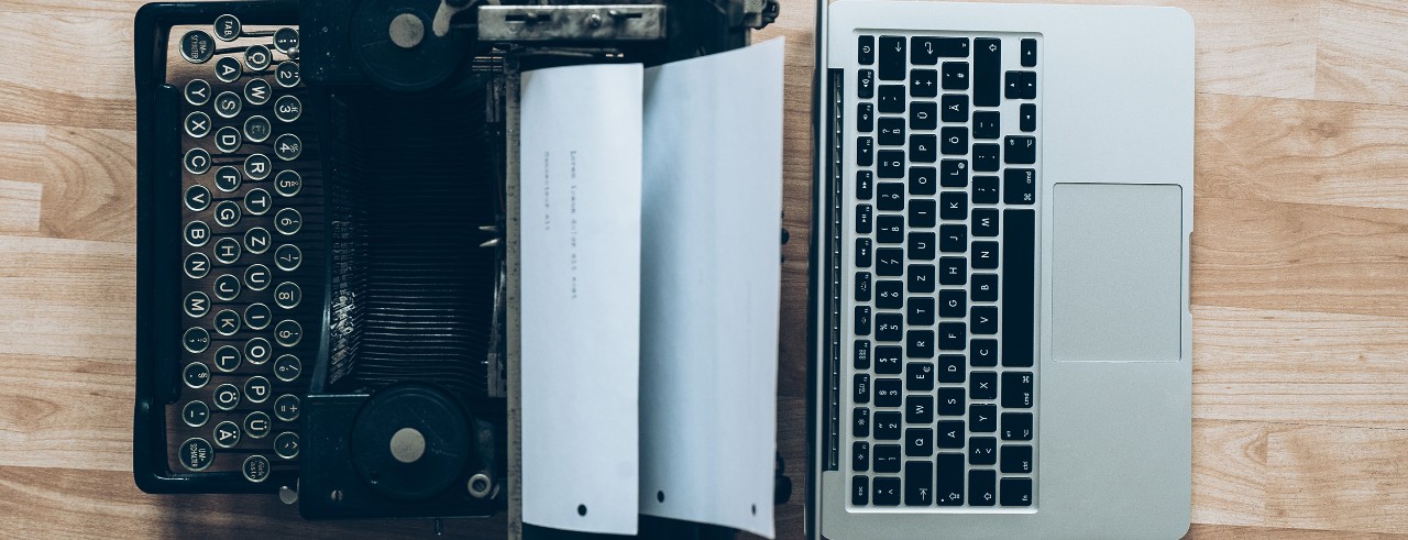 An old typewriter and a new laptop with the screens back to back, bird's eye view.