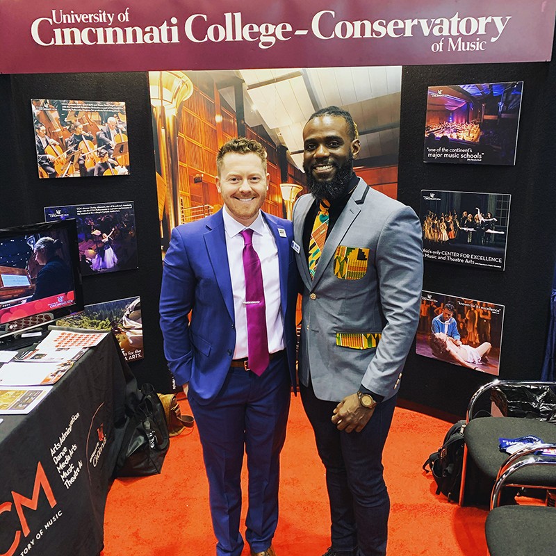 Two men pose in from of an information booth at a conference.