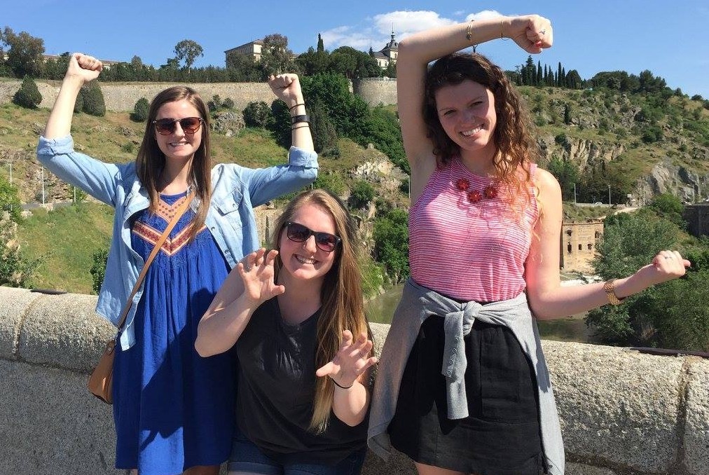 Grace Hertlein poses with two other students in front of a grassy hill in Spain, two with their arms up to form the letters "UC" while the other bares her teeth like a bearcat.