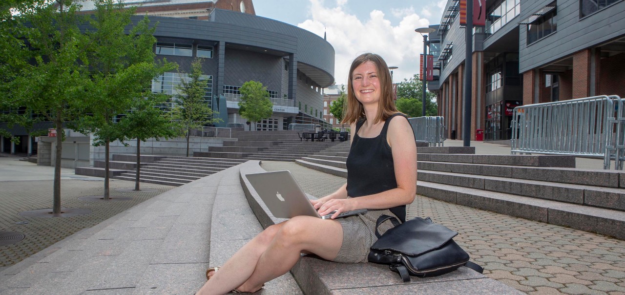 Recent UC grad Ravenna Rutledge sits with laptop along UC's MainStreet.