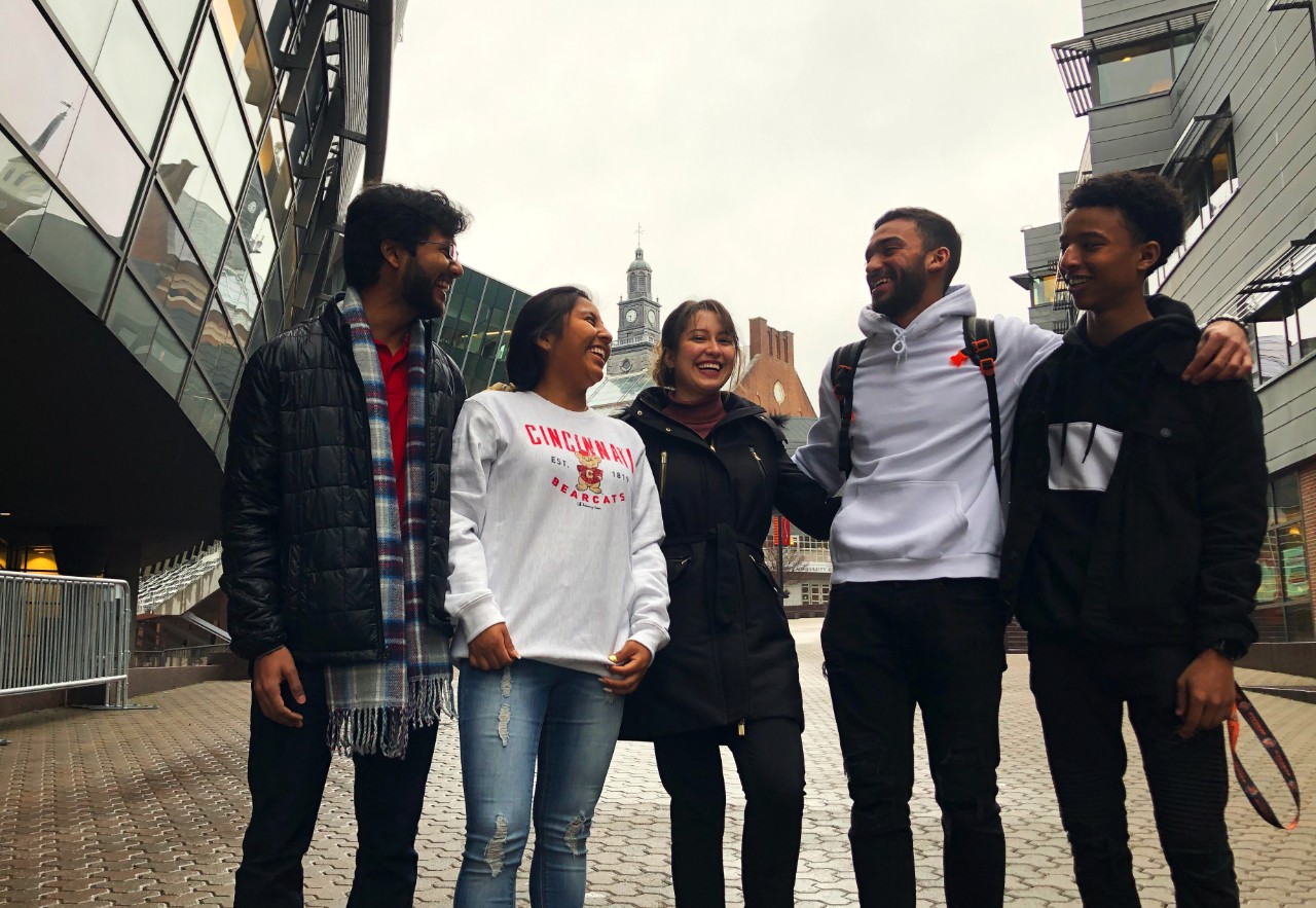 A group of students pose, smiling, on a paved walkway with UC buildings in a wide variety of architectural styles behind them