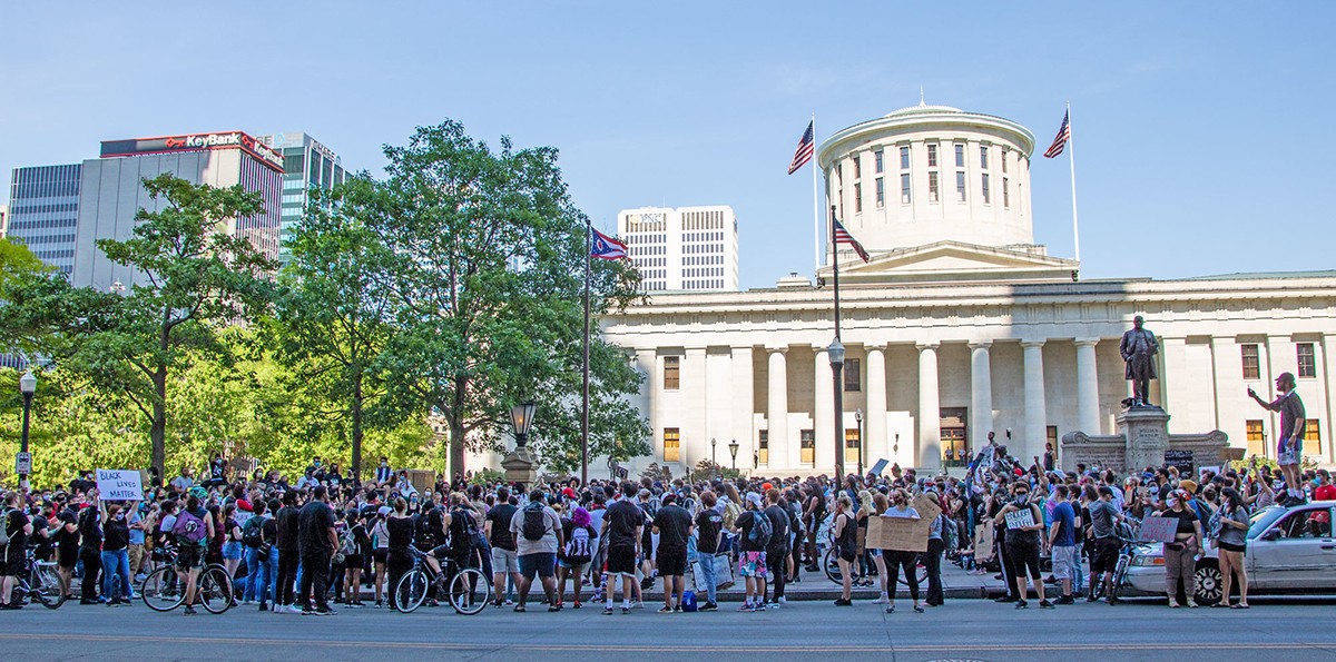 protesters gather in front of a large white statehouse