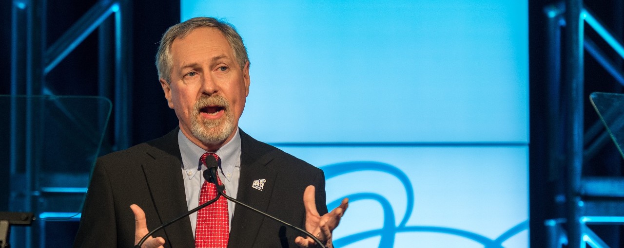 gentleman with suit jacket, dress shirt and red tie speaks at a microphone with his hands open in conversation while standing at a clear podium in front of a light blue and dark blue background on a stage