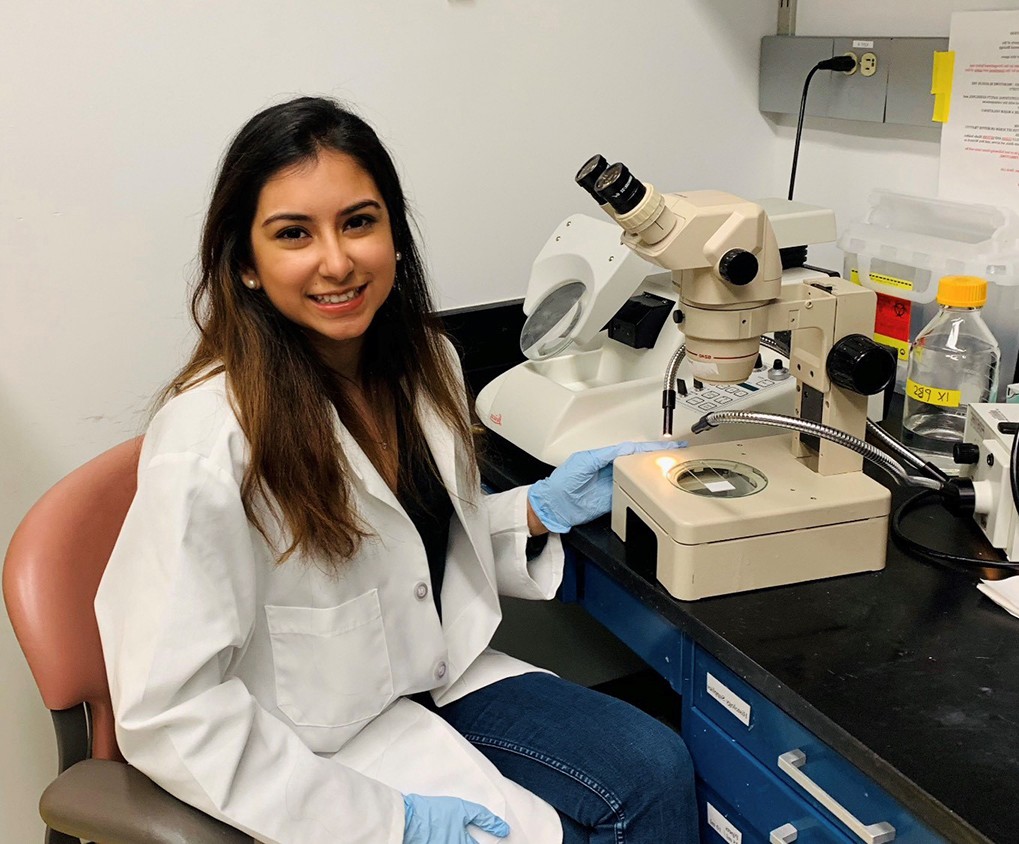 Trichy, a young woman in a lab coat and gloves, sits in front of a microscope at a lab bench.