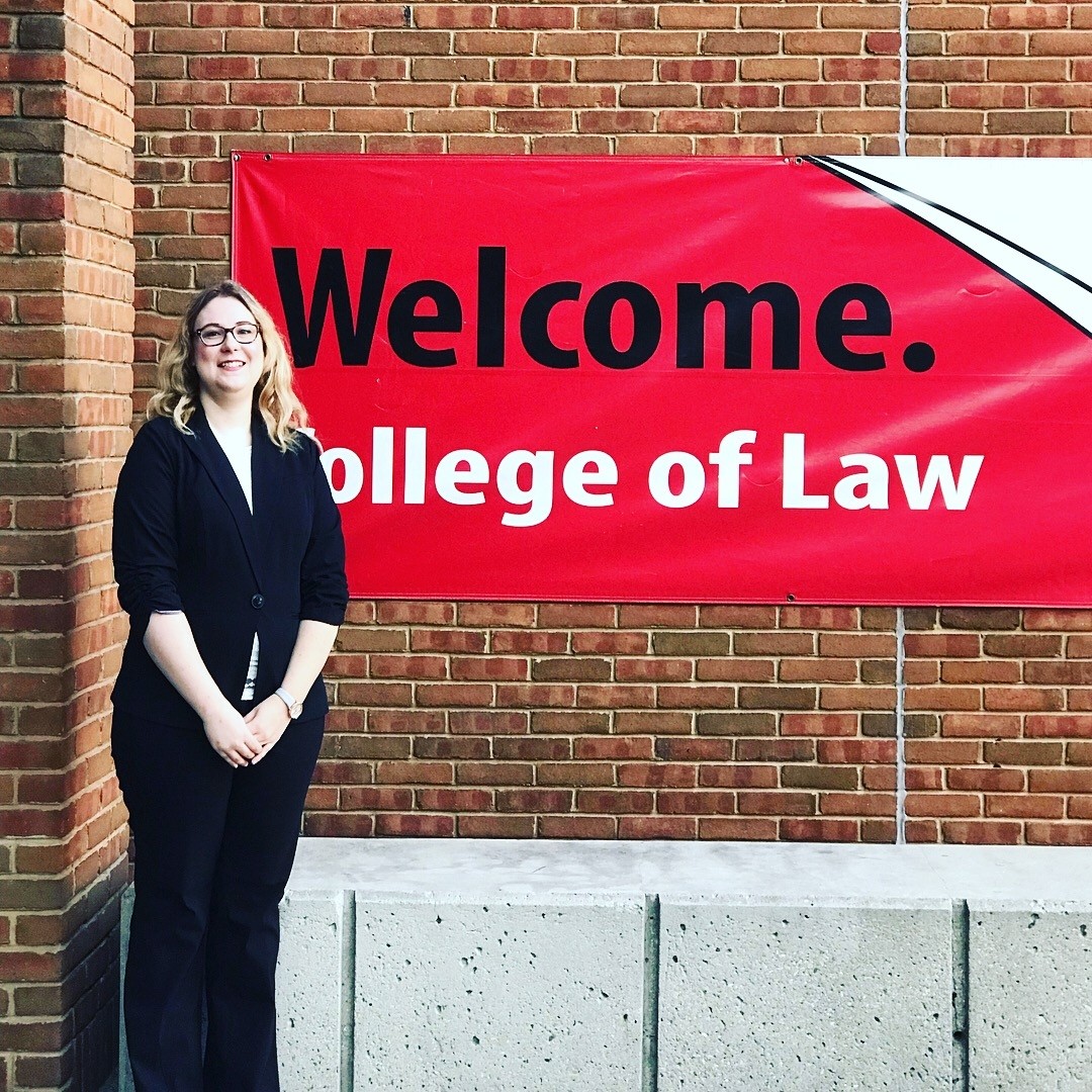 A woman standing outside a building with a sign that reads, "Welcome. College of Law."