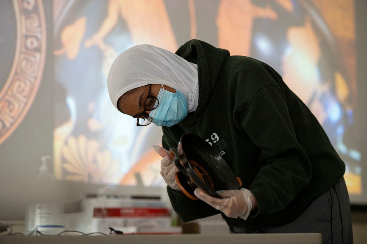 A student wearing a face mask and gloves leans over to closely inspect a clay cup in gloved hands.