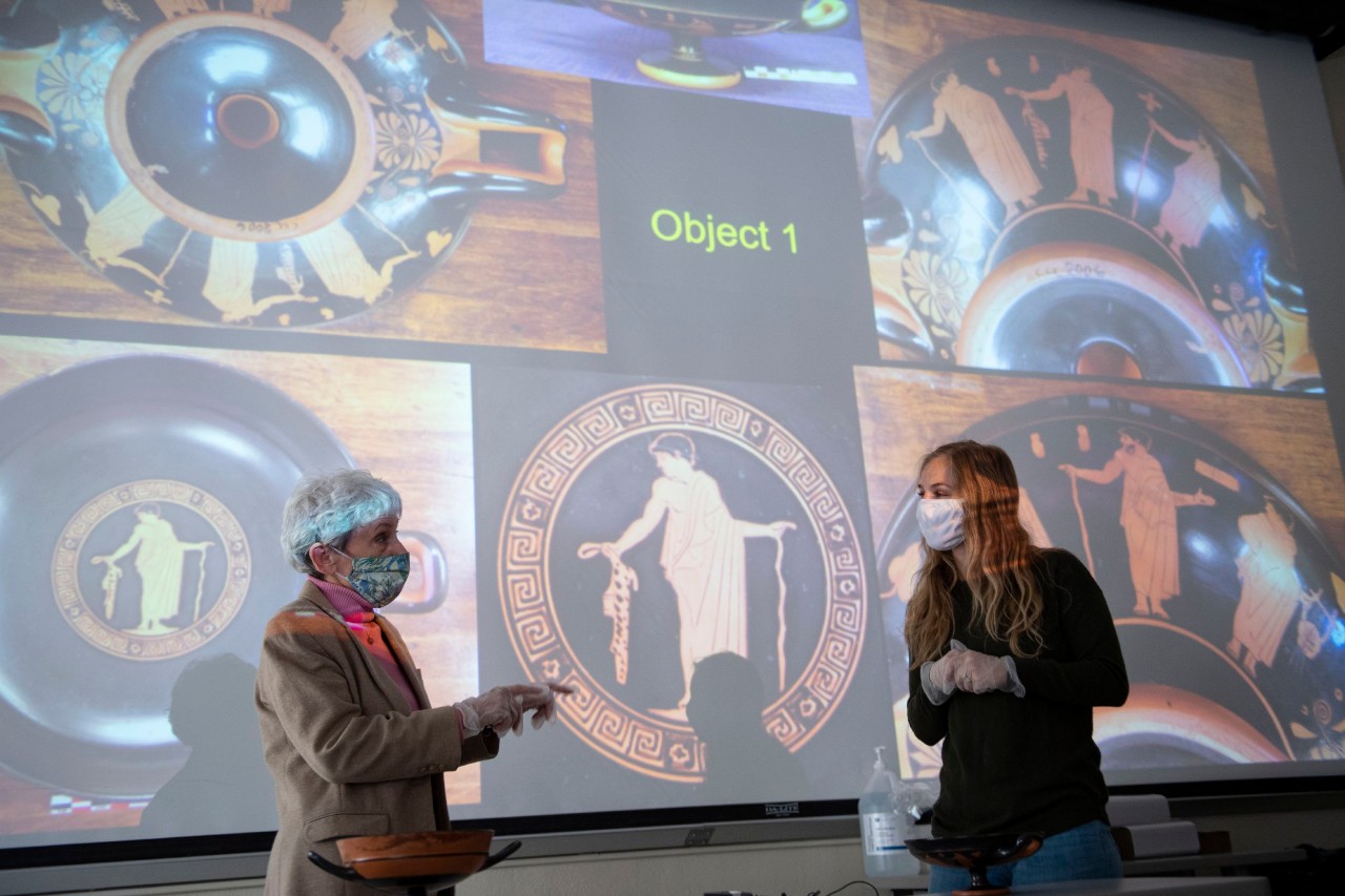 Two people wearing face masks talk in front of a projected image showing photos of an ancient Greek cup in a lecture hall.