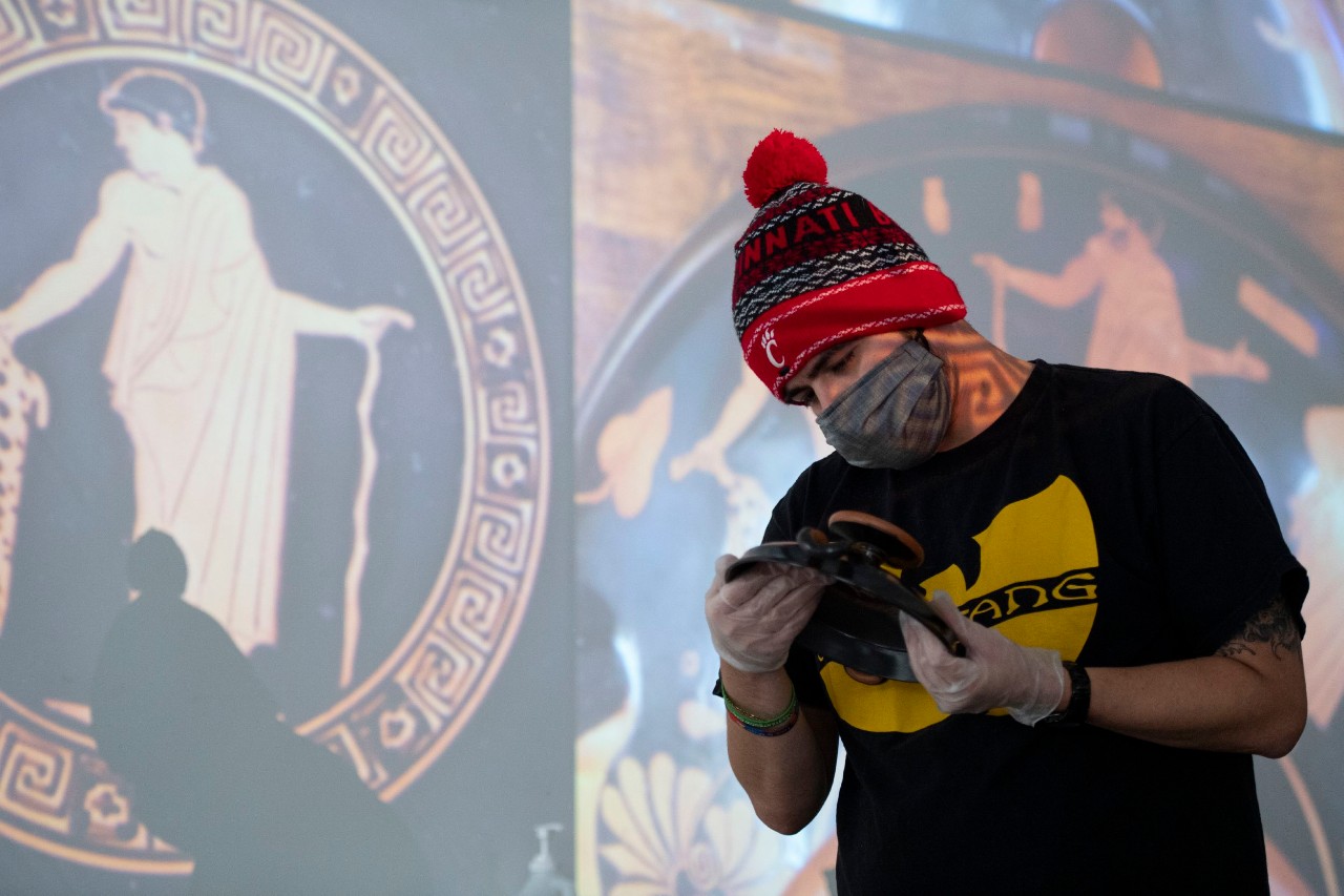 A student wearing a face mask and gloves holds an ancient clay cup in front of a projected image of the figures on the cup.