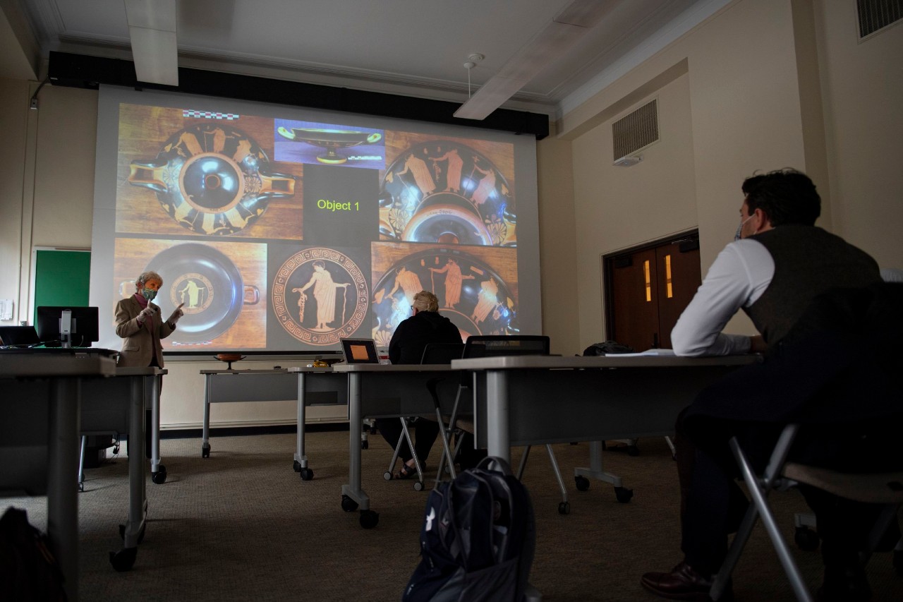 A professor stands next to projected images of clay artwork in a lecture hall.