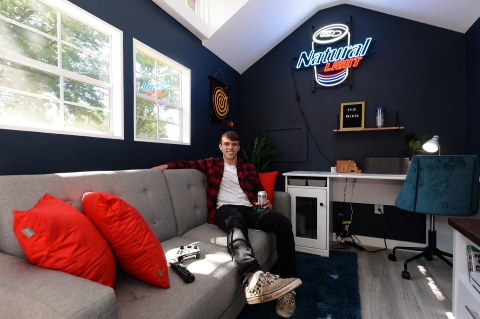 A male college student sitting on a couch inside of a mobile home with a neon Natural Light sign hung on the rear wall.
