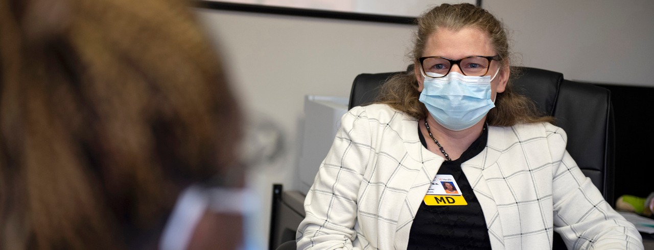 a female doctor wearing a mask listening to a patient while sitting in a chair