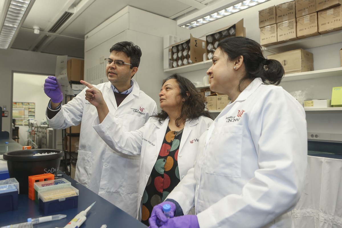 A man and two women in lab coats point and analyze a specimen the man holds up