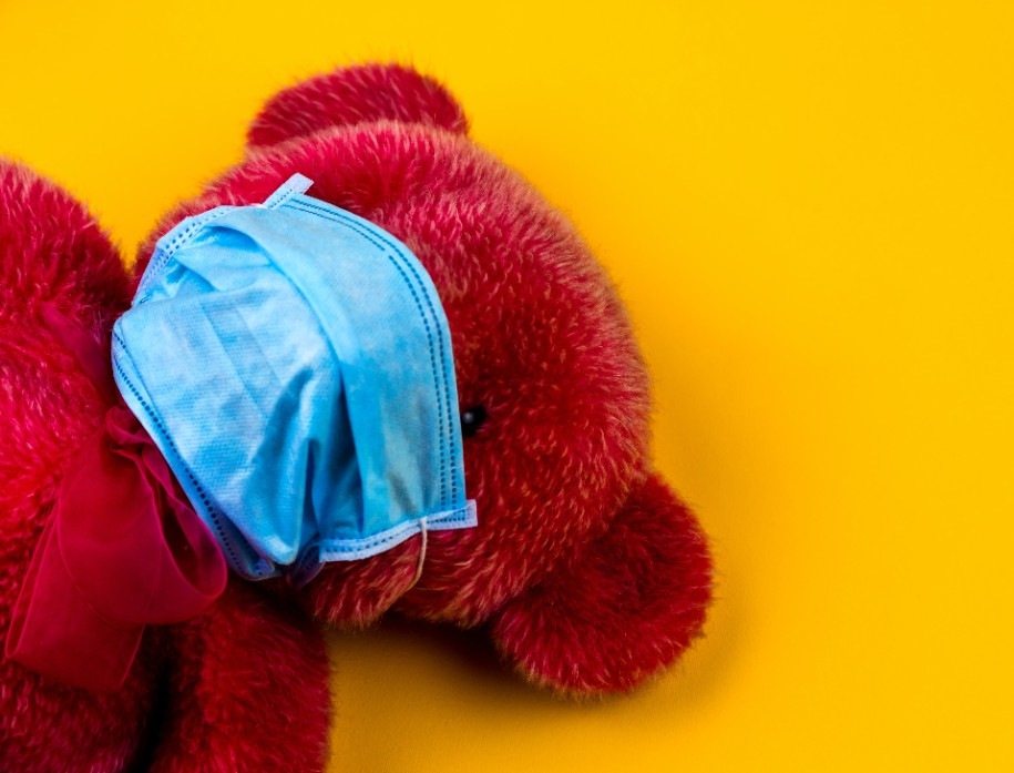 A teddy bear wears a surgical mask against a yellow backdrop.
