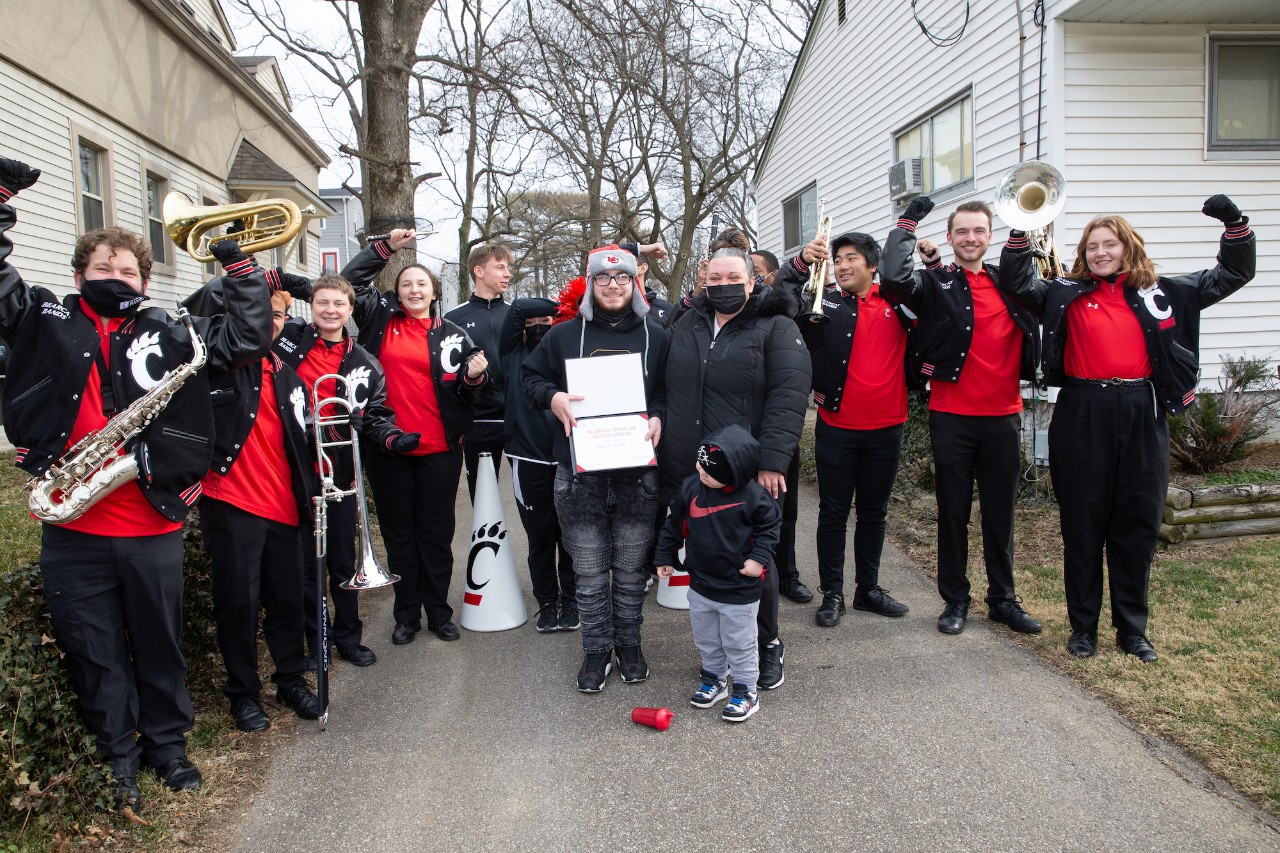 Marcus Elliott standing outside with the UC band and UC officials. Elliott is holding a certificate with his Marian Spencer Scholarship.