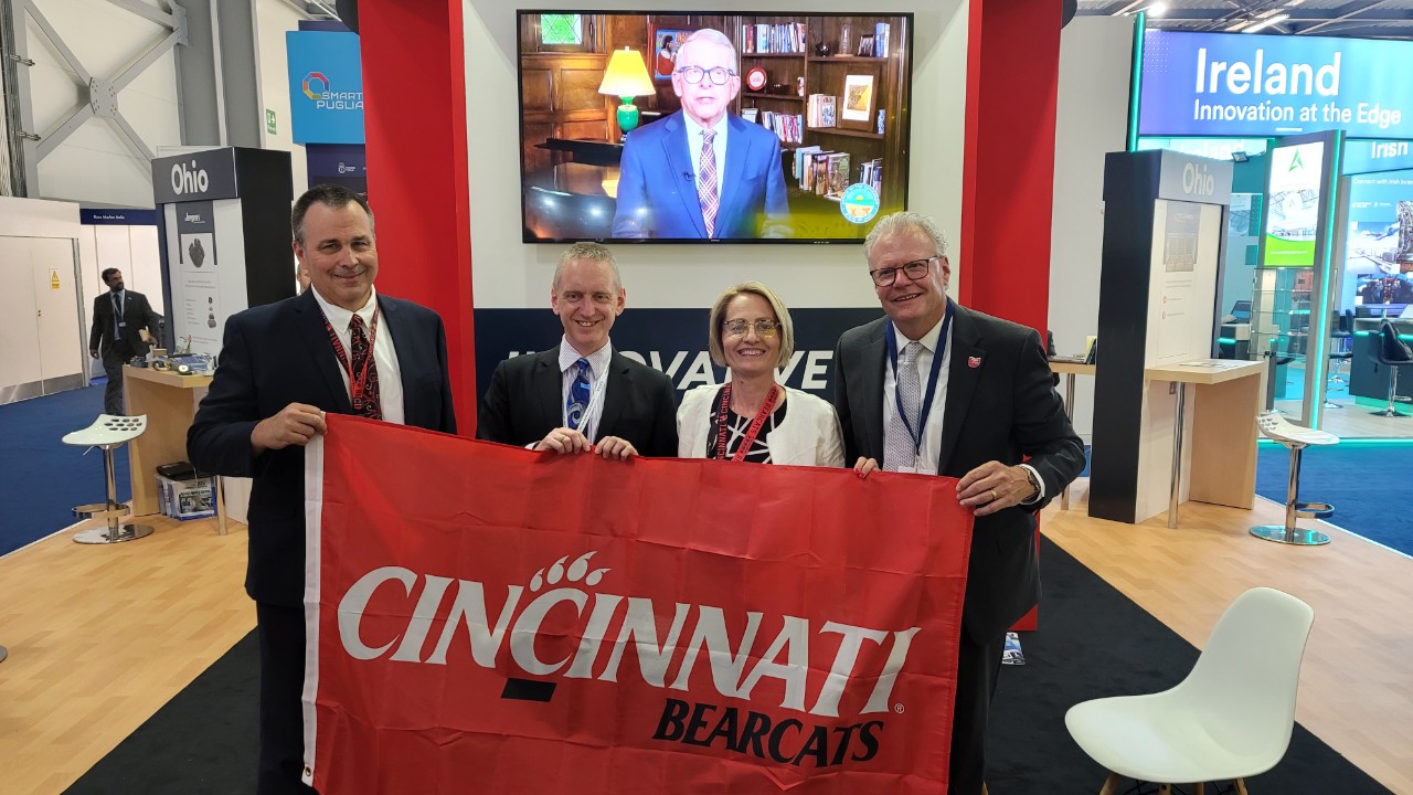 three men and one woman wearing suits pose while holding a red Cincinnati Bearcats flag