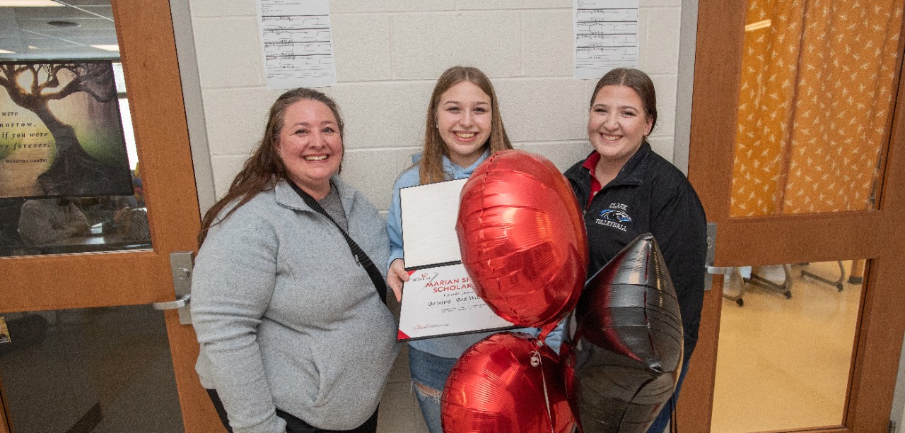 Brooke Bartholomew holds balloons and a certificate while celebrating her scholarship announcement with her family in a school hallway.