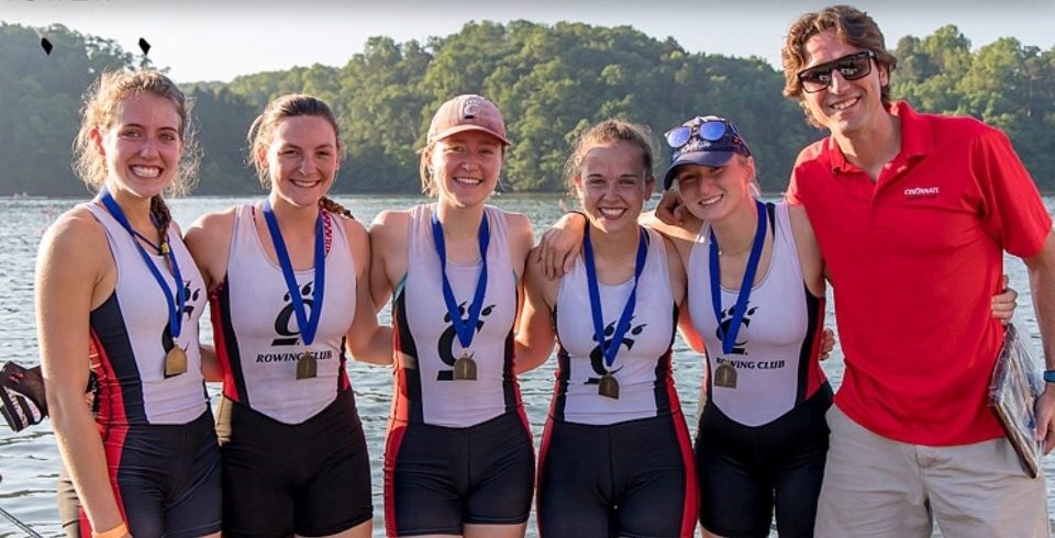 Rowing team and coach stands with smiling with medals around their necks in front of water