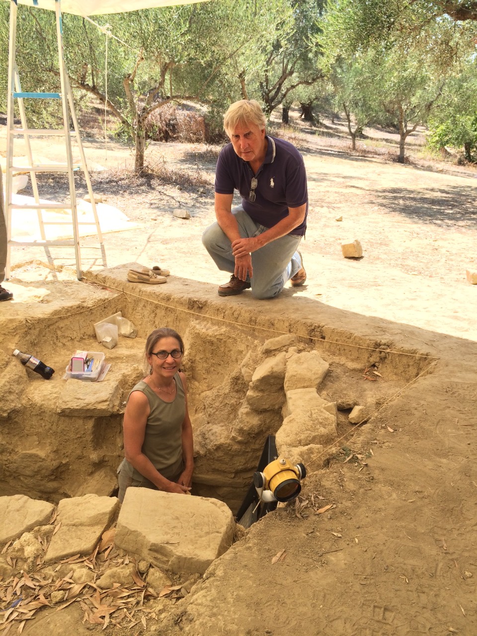 Jack Davis kneels at the edge of an excavation in which Sharon Stocker is standing in an olive grove shaded from the bright sun by a tarpaulin.