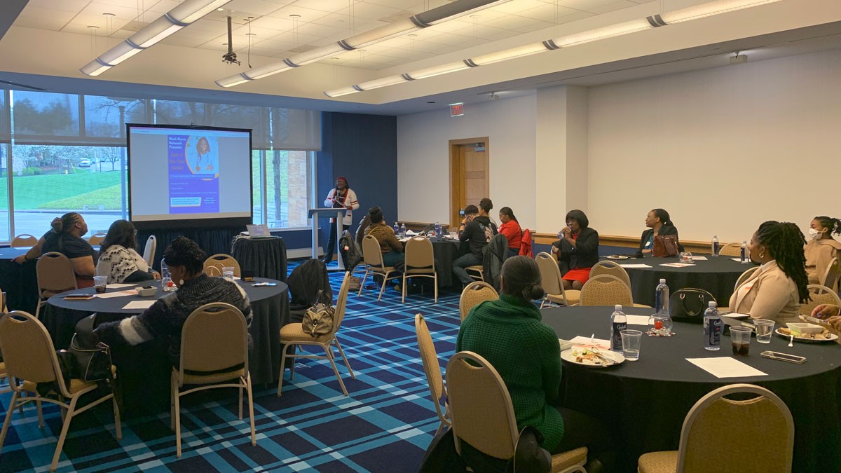 Karen Bankston stands at a podium in front of Black Nurse Network participants at round tables around the room.