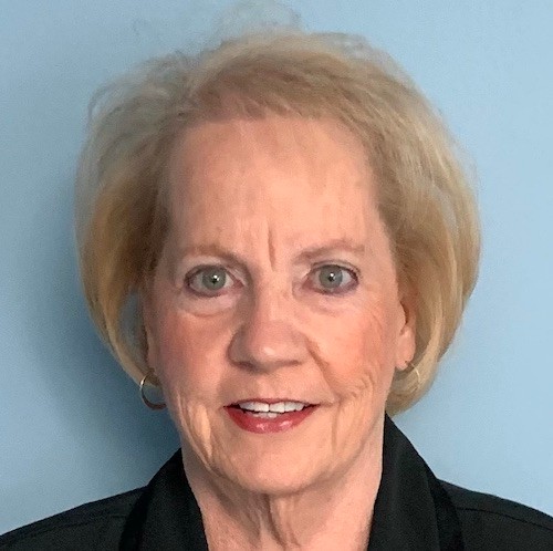 woman with short blonde hair and black top standing and smiling in front of light blue background