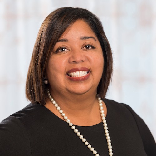 black woman with chin length hair and pearl necklace and black top smiles for a headshot against a light grey background