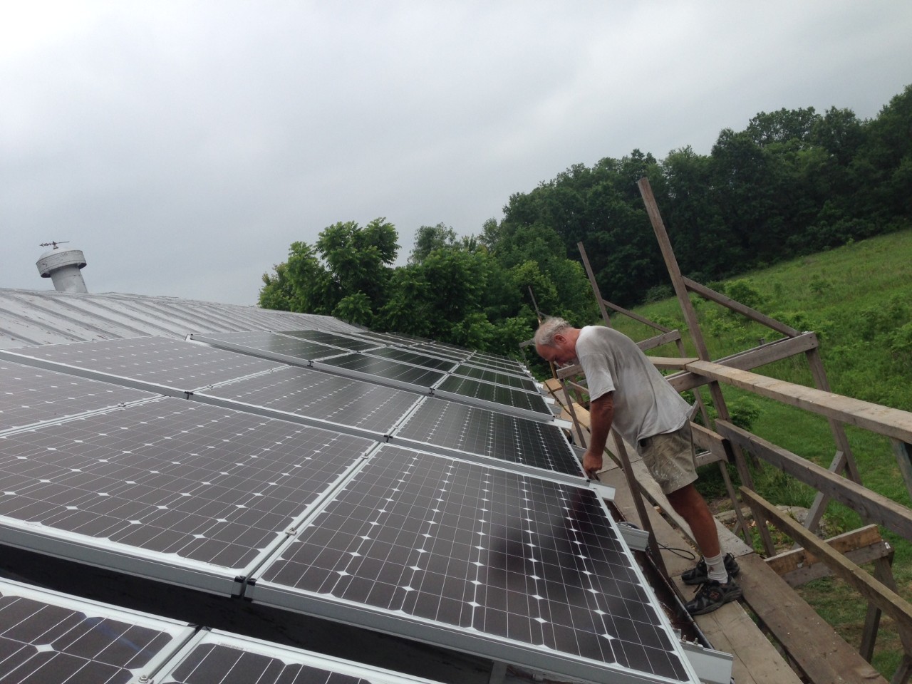 A man stands on a scaffold installing solar panels on a high barn roof surrounded by woods.