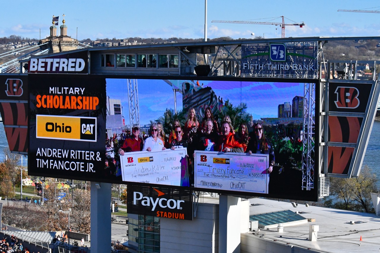 A Jumbotron image at Paycor Stadium shows two students holding up oversized checks surrounded by supporters. 