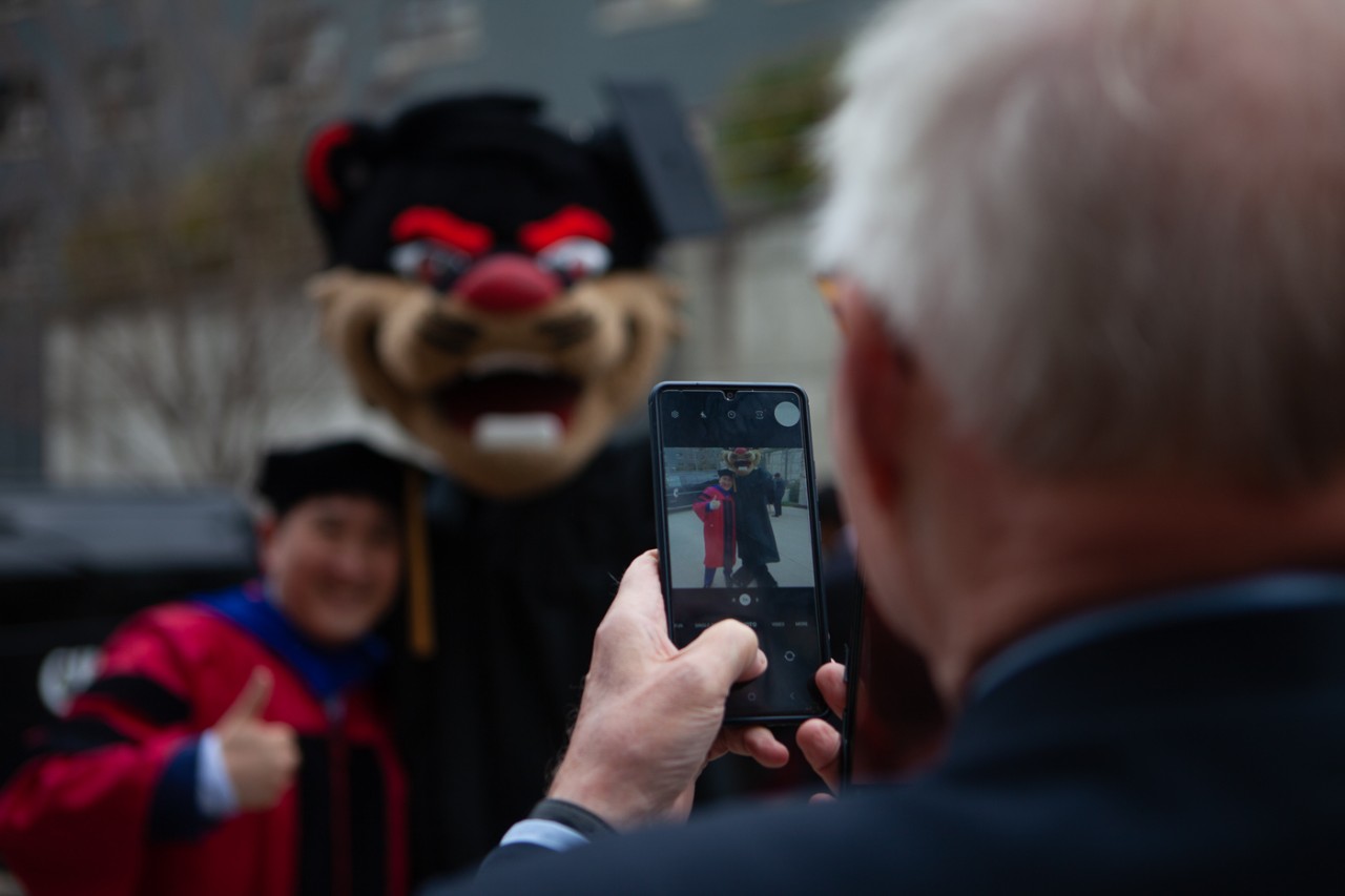 A person takes a cell phone photo of the Bearcat mascot in a cap and gown with a grad wearing a cap and gown.