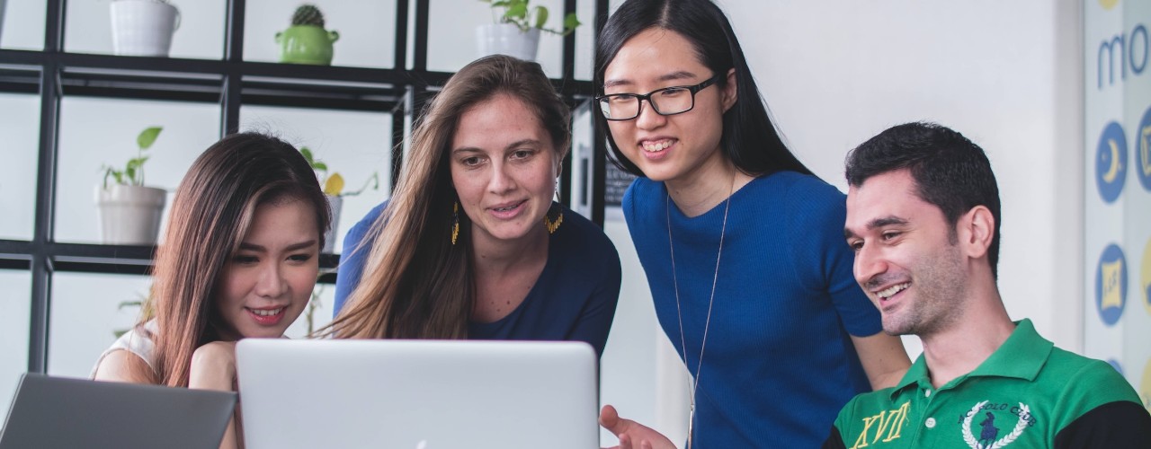 Three women and one man sit at a laptop smiling.