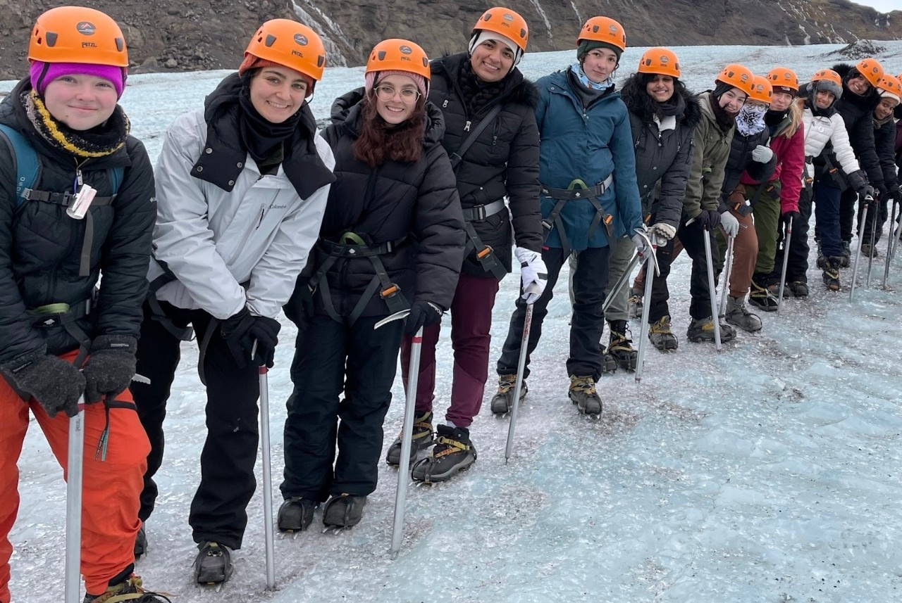 Students in hard hats stand in a line and smile while hiking on an Icelandic glacier.