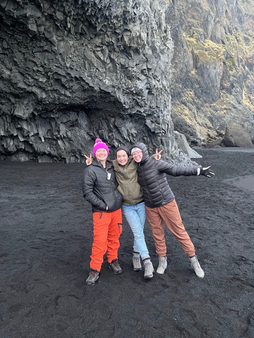Three women, dressed in heavy winter coats, stand on a black sand beach with a rocky cliff in the background.