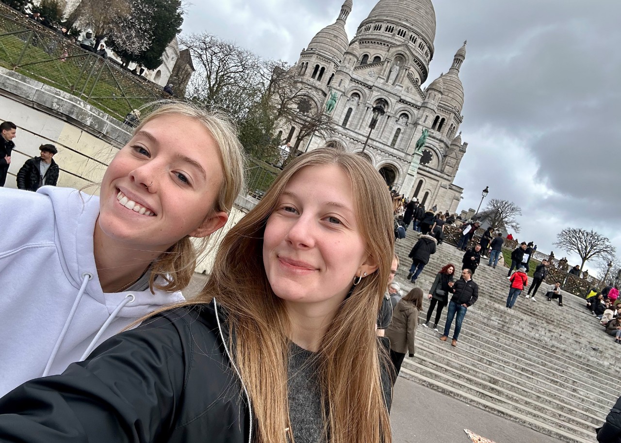 Two young women take a selfie in a plaza before a long, wide flight of steps; in the background behind them, the Sacre Coeur basilica in Paris is visible.